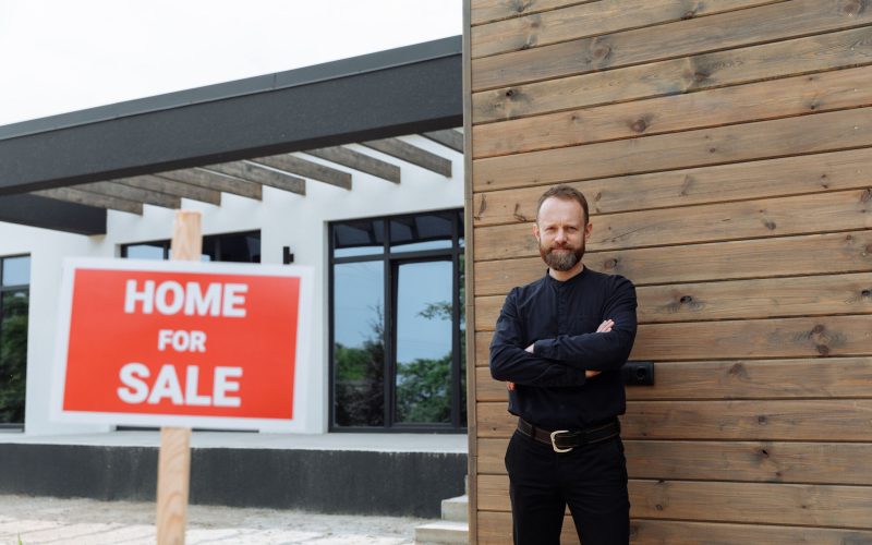 Real estate agent standing by a modern home with a 'Home for Sale' sign.